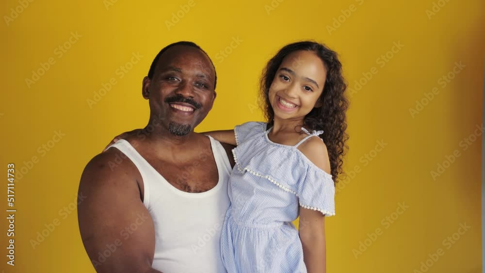 African man holding his daughter in his arms. His daughter kisses him on the nose and they smile for the camera. Black man with his family stands against a yellow background. African man holding his daughter in his arms. His daughter kisses him on the nose and they smile for the camera. Black man with his family stands against a yellow background.