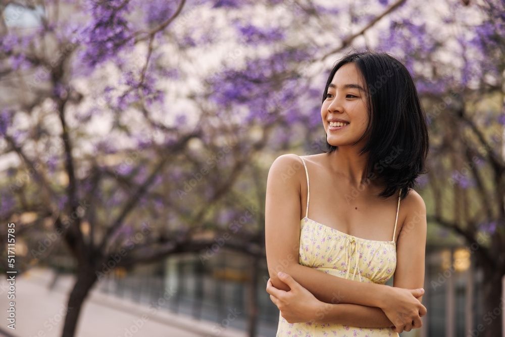Attractive charming brunette woman looking with smile away . Chinese young lady in yellow dress at the park keeps arms crossed. Street style concept Attractive charming brunette woman looking with smile away . Chinese young lady in yellow dress at the park keeps arms crossed. Street style concept