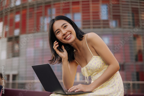 Chinese cheerful happy woman holding smartphone and laptop on legs . Dark-haired young lady luffing look away sitting in the city. Concept of emotional, lifestyle mood
