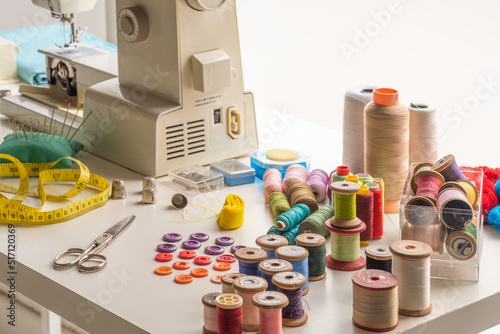 work table with spools of thread or bobbins. next to it, scissors and a tape measure, in the background, a sewing machine
 photo