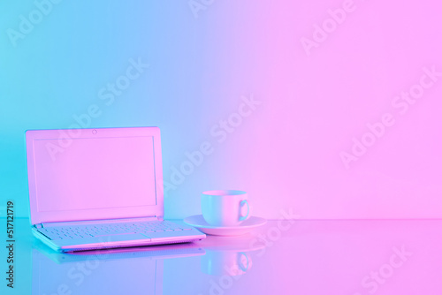 Office table with notebook computer and cup of coffee in vibrant gradient holographic neon colors with copy space. Concept art. Minimal office surrealism.