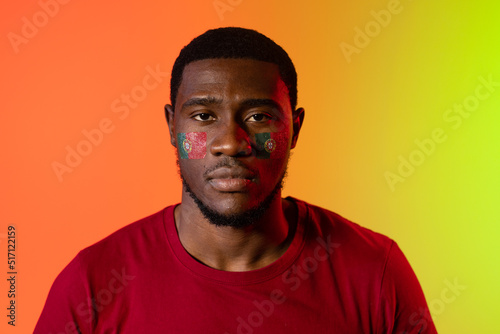 Portrait of african american male supporter with flag of portugal on cheeks over orange lighting