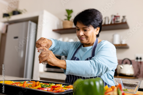 Biracial mature woman with short hair sprinkling pepper with shaker on vegetables in tray in kitchen