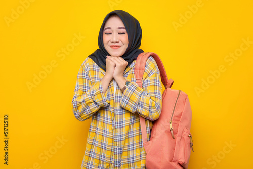 Smiling young Asian Muslim woman student in casual clothes and backpack, making a praying gesture with closed eyes isolated on yellow background. Education school university college concept © Bangun Stock Photo