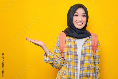 Smiling young Asian Muslim woman student in casual clothes and backpack, showing copy space on palm isolated on yellow background.Education school university college concept