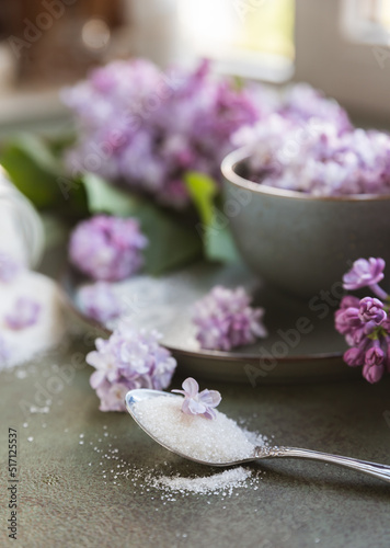 Flavored sugar with lilac flowers in glass jar and branch of beautiful lilac, green concrete background. Edible flowers in cooking and confectionery. Flavoring ingredients.