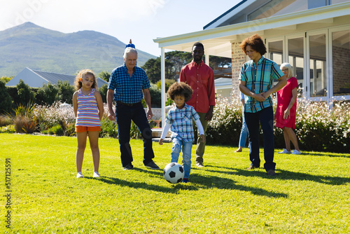 Multiracial multigeneration family looking at boy playing soccer on grassy field in yard in summer