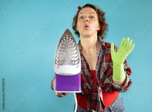 An adult pin-up woman shows an iron at the camera on a blue background. Portrait of a woman in a plaid shirt. Iron clothes photo