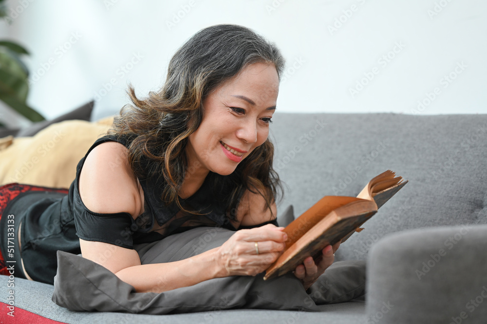 Obraz premium Positive middle aged female resting on couch and reading favorite literature, enjoying leisure weekend time at home