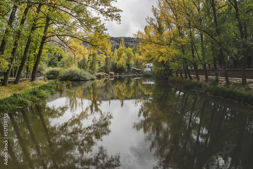 Autumn landscape in Soria with yellow and green trees reflected in the Duero river with some white clouds also reflecting in the water.