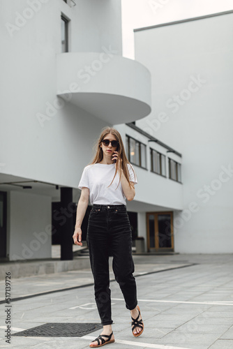 Young woman in white t-shirt standing in front of grey building and using smartphone. Urban style and street fashion. Girl in sunglasses with phone. Chatting, messaging, reading