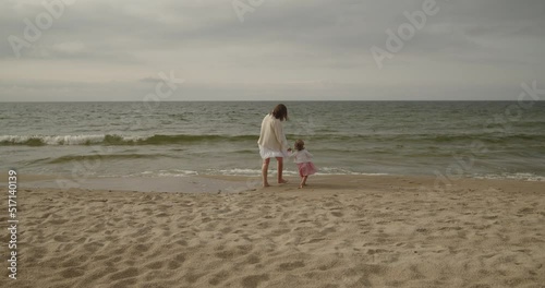 Young mother and baby daughter spend time on the beach together. Mom and toddler cute little girl walk by the sea alone holding hands. Motherhood, maternity, single mother, family concept.