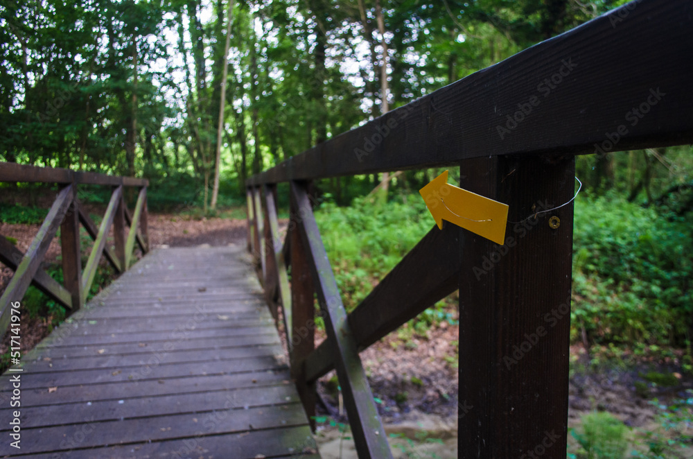 La freccia gialla che indica la direzione della Via Postumia, cammino che parte da Aquileia e arriva a Genova, inchiodata sul parapetto di un ponte di legno che attraversa un ruscello La freccia gialla che indica la direzione della Via Postumia, cammino che parte da Aquileia e arriva a Genova, inchiodata sul parapetto di un ponte di legno che attraversa un ruscello