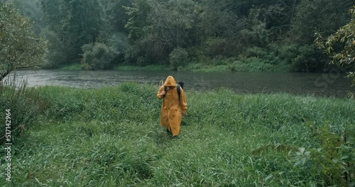 Heavy rain, hiker in a yellow raincoat with a backpack in the tall grass next to the river. The concept of backpacking, lifestyle, hike, extreme hobbies and travel