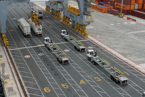 Tracks or lorries in lines with numbers are waiting below gantry cranes for loading containers from the ship in container terminal. on the left are stowed boxes.