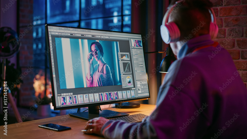 Young Creative Man in Headphones Using Computer in Stylish Loft Apartment in the Evening. Graphic Designer Working from Home, Editing Fashion Photo for Online Store. Urban City View from Big Window. Young Creative Man in Headphones Using Computer in Stylish Loft Apartment in the Evening. Graphic Designer Working from Home, Editing Fashion Photo for Online Store. Urban City View from Big Window.