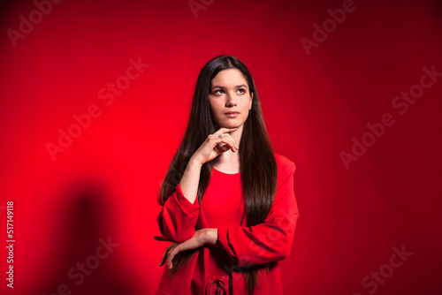 Portrait cute thinking teen girl wearing stylish red costume over red background wall looking away