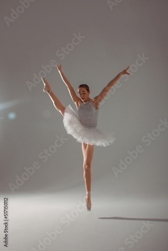 classic ballet trick. young gymnast dancer, fit girl ballerina is jumping in white ballet tutu like swan in cross twine in air near a white wall background in studio light. ballet concept, free space