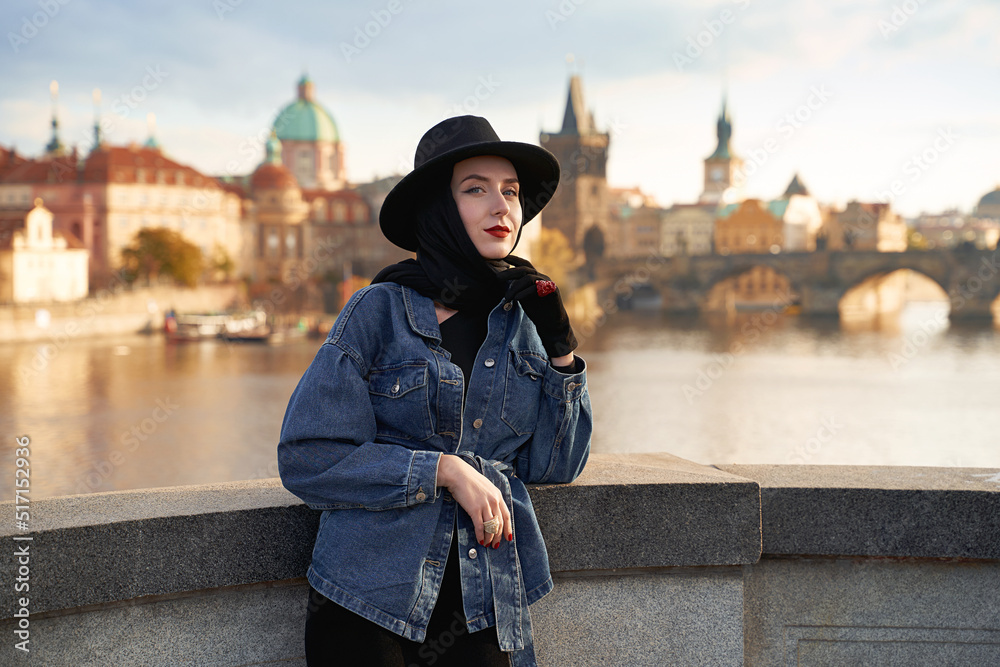 Stylish beautiful young woman earing black hat in Prague with Charles Bridge on background. Elegant retro lady fine art portrait.