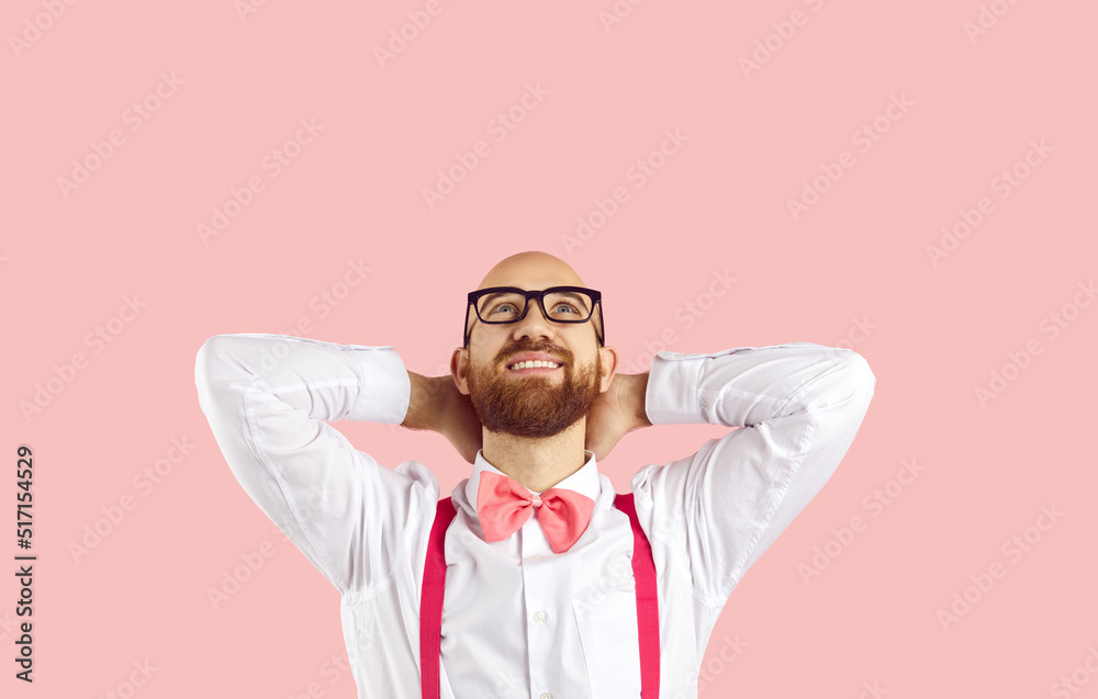 Young happy man holds his hands behind his head and dreamily looks up on pastel pink background. Caucasian handsome stylish man in white shirt, bow tie and suspenders dreams or imagines. Copy space. Young happy man holds his hands behind his head and dreamily looks up on pastel pink background. Caucasian handsome stylish man in white shirt, bow tie and suspenders dreams or imagines. Copy space.