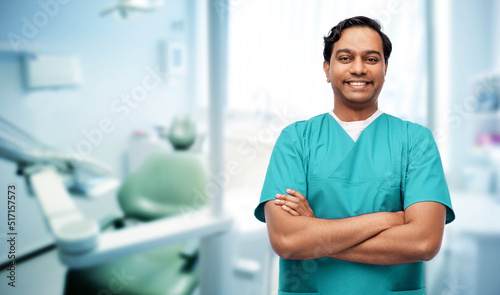 healthcare, profession and medicine concept - happy smiling doctor, dentist or male nurse in blue uniform with crossed arms over medical office at dental clinic on background
