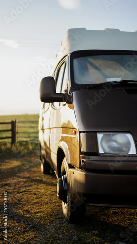Shot of a brown and white retro fashioned camper van laying near the wooden fence of a green field during the sun rise moments of the road trip. © White