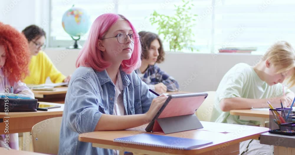 Portrait of young pupil with happy facial expression sitting at table with wireless tablet in modern classroom. Pink haired beautiful child looking at camera with smile. Portrait of young pupil with happy facial expression sitting at table with wireless tablet in modern classroom. Pink haired beautiful child looking at camera with smile.