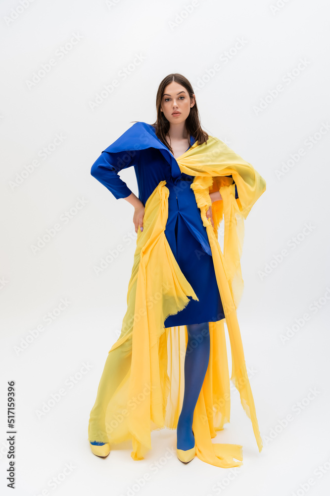 full length of confident ukrainian woman in blue and yellow outfit posing with hands on hips on grey.