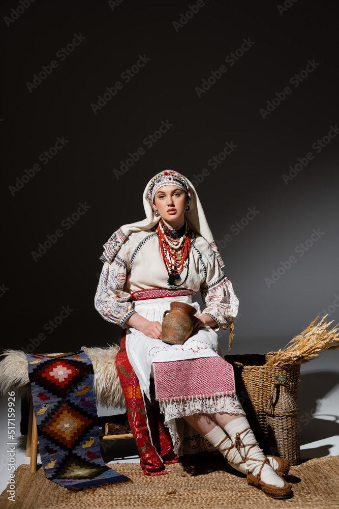 full length of young ukrainian woman in traditional clothing with ornament holding clay pot while sitting on bench on black.