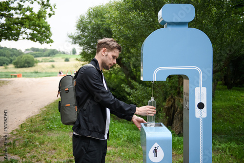Man refilling his water bottle at the city. Free public water bottle refill station. Sustainable and green city. Male in black coat. Tap water to reduce plastic bottle usage. Drinking water dispenser