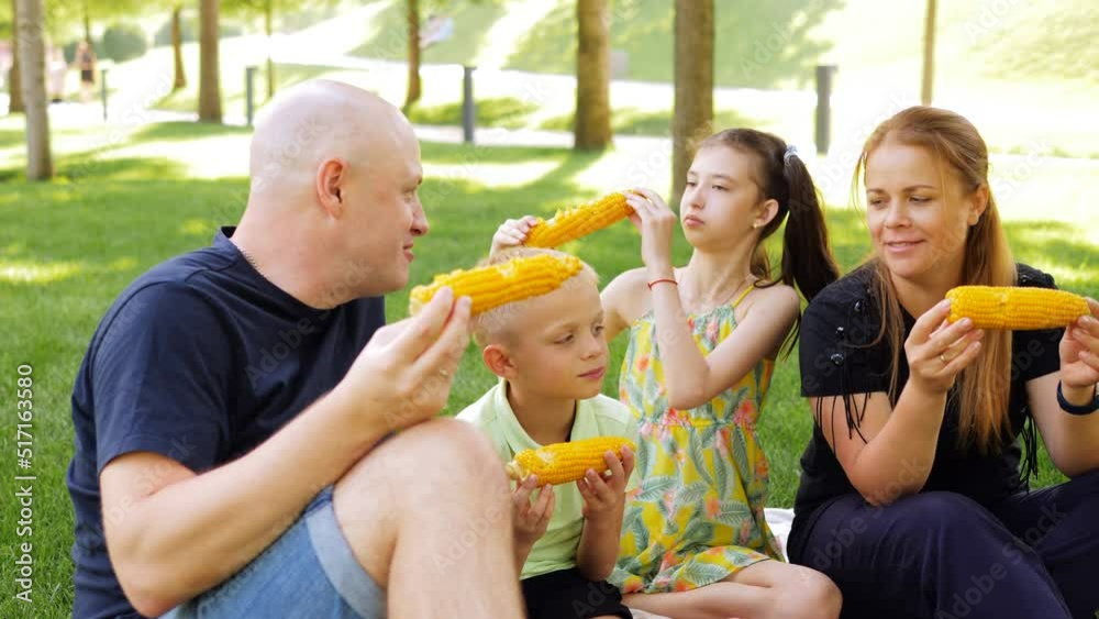 A man and a woman with two children on a picnic in the park are eating corn cobs with an appetite. A man and a woman with two children on a picnic in the park are eating corn cobs with an appetite.