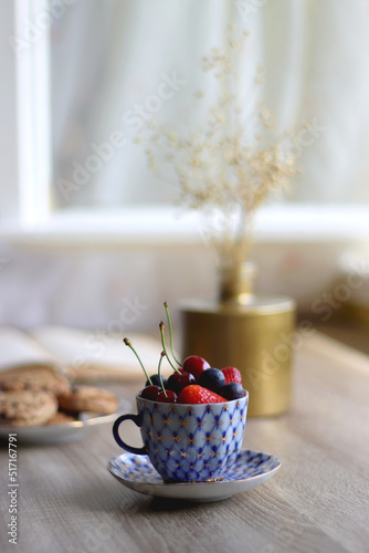 Plate of chocolate chip cookies, cup filled with strawberries, blueberries and cherries, open book and vase with gypsophila flowers on the table. Selective focus.