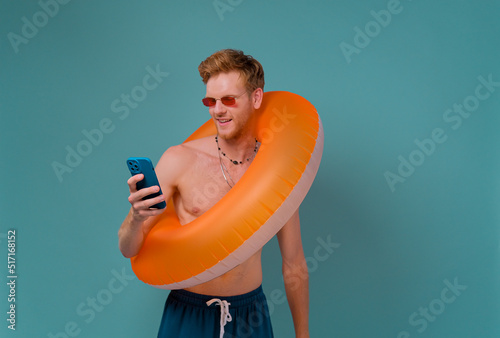 Red-haired man with orange rubber ring and smartphoneusing an app on a smartphone. Summer vacation concept isolated on blue background. photo