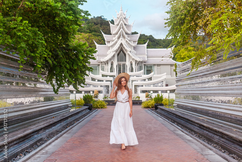 Traveler woman in white dress walking in front of a beautiful white temple in thai style. Travel and vacation in Asia. Young woman in hat near beautiful buddhist temple on Phuket Island in Thailand