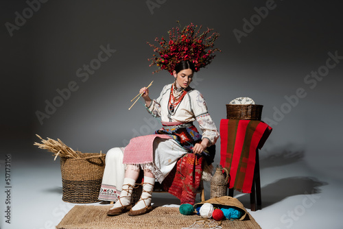 full length of young ukrainian woman in traditional clothes and red wreath with flowers holding knitting needles on dark grey. photo