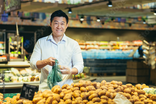 Portrait of an Asian shopper in the grocery department of a supermarket, a man chooses vegetables and buys potatoes, smiling and looking at the camera