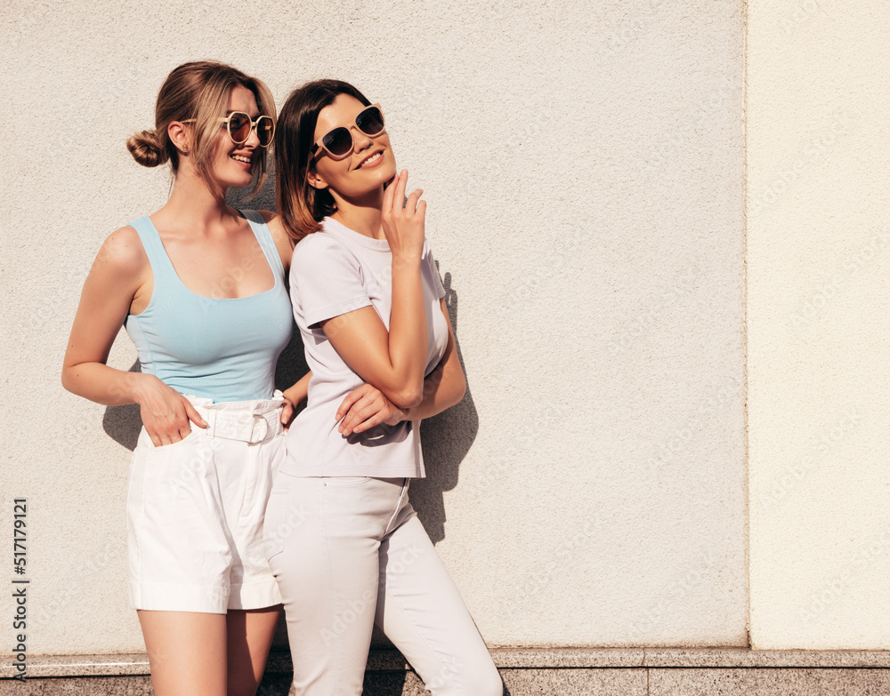 Portrait of two young beautiful smiling hipster female in trendy summer white t-shirt clothes.Sexy carefree women posing on street background. Positive models having fun, hugging. In sunglasses