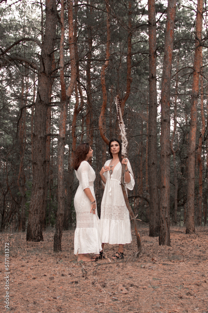 female witch cosplay. two brunette girls in white boho dresses are standing close with high wooden witch staff in hand and looking apart on pine forest background. lifestyle shaman concept, free space female witch cosplay. two brunette girls in white boho dresses are standing close with high wooden witch staff in hand and looking apart on pine forest background. lifestyle shaman concept, free space