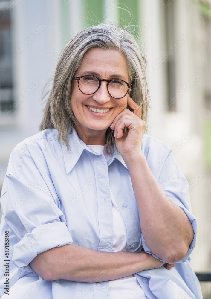 Portrait of beautiful mature grey hair woman wearing glasses standing outdoor enjoying free time vacation traveling around world at retirement. Mature woman with perfect skin wearing blue shirt Portrait of beautiful mature grey hair woman wearing glasses standing outdoor enjoying free time vacation traveling around world at retirement. Mature woman with perfect skin wearing blue shirt