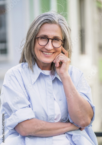 Portrait of beautiful mature grey hair woman wearing glasses standing outdoor enjoying free time vacation traveling around world at retirement. Mature woman with perfect skin wearing blue shirt