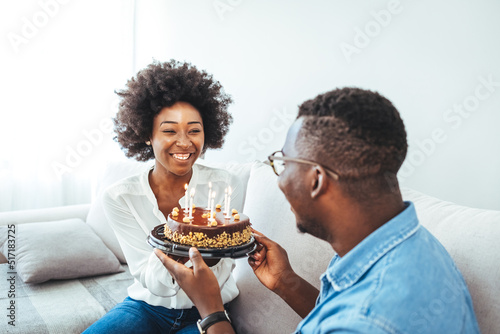 Young positive wife making surprise for husband  excited man holding gift box and being so happy while sitting in Living room at table with cake