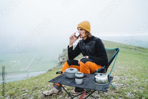 Drink hot delicious coffee sitting on a chair in the mountains, camping kitchen, guy tourist relaxing in the mountains enjoying a beautiful view, vacation in nature, cloudy weather in the mountains