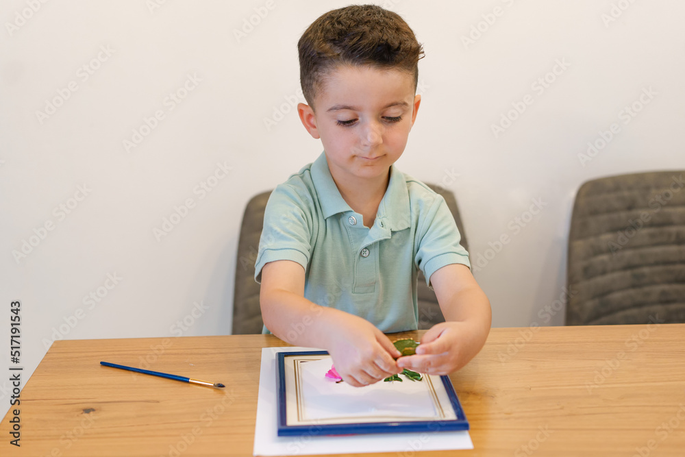 Child with album, leaves, and flowers making herbarium on dining wood table in home. Cute little boy arranging pressed flowers onto a picture frame. Child with album, leaves, and flowers making herbarium on dining wood table in home. Cute little boy arranging pressed flowers onto a picture frame.