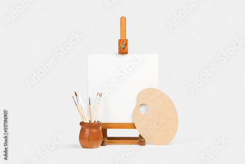Lienzo en blanco sobre un caballete de madera junto a una paleta de pintura y pinceles sobre un fondo gris claro. Vista de frente y de cerca. Copy space photo