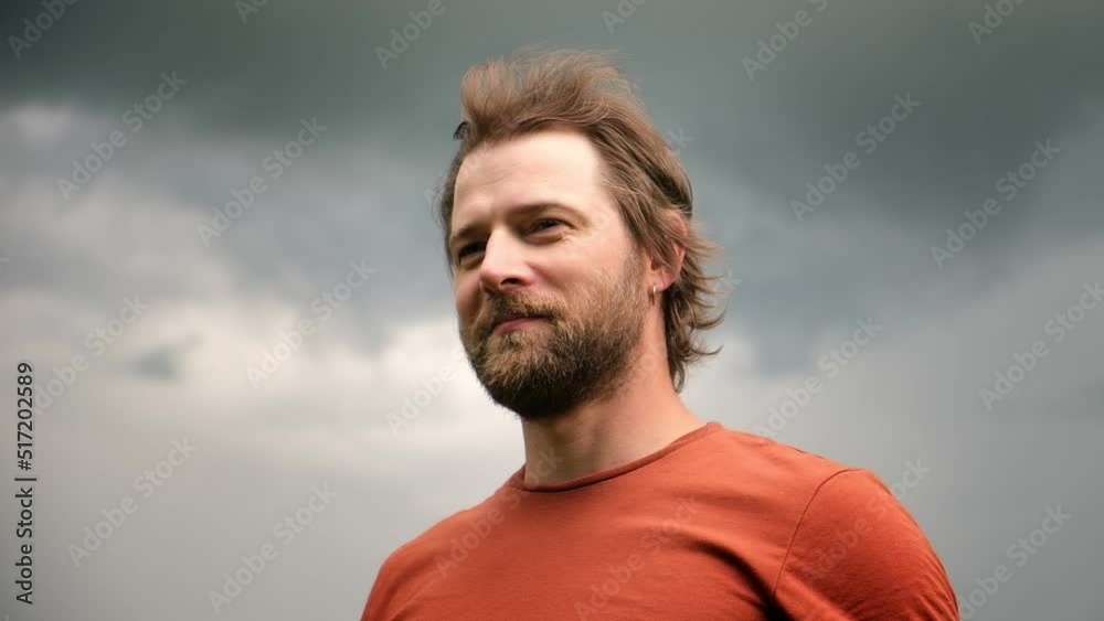 Portrait of handsome young man in orange t-shirt outdoors. Male with long hair and beard feel happy, calm and serious. Dramatic dark sky background. Man looking away into distance. Diversity People. Portrait of handsome young man in orange t-shirt outdoors. Male with long hair and beard feel happy, calm and serious. Dramatic dark sky background. Man looking away into distance. Diversity People.