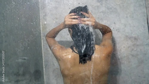 Back view of naked woman washing hair with shampoo while taking a shower