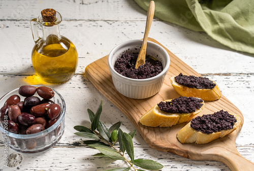 Olive pate in bowl and spread on bread slices on wooden cutting board.  photo