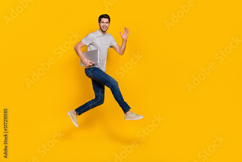 Full body profile photo of cool brunet millennial guy run with laptop wear t-shirt jeans footwear isolated on yellow background