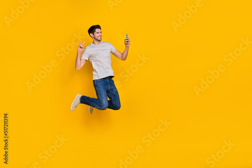 Full body photo of cute brunet young guy jump do selfie wear t-shirt jeans footwear isolated on yellow background
