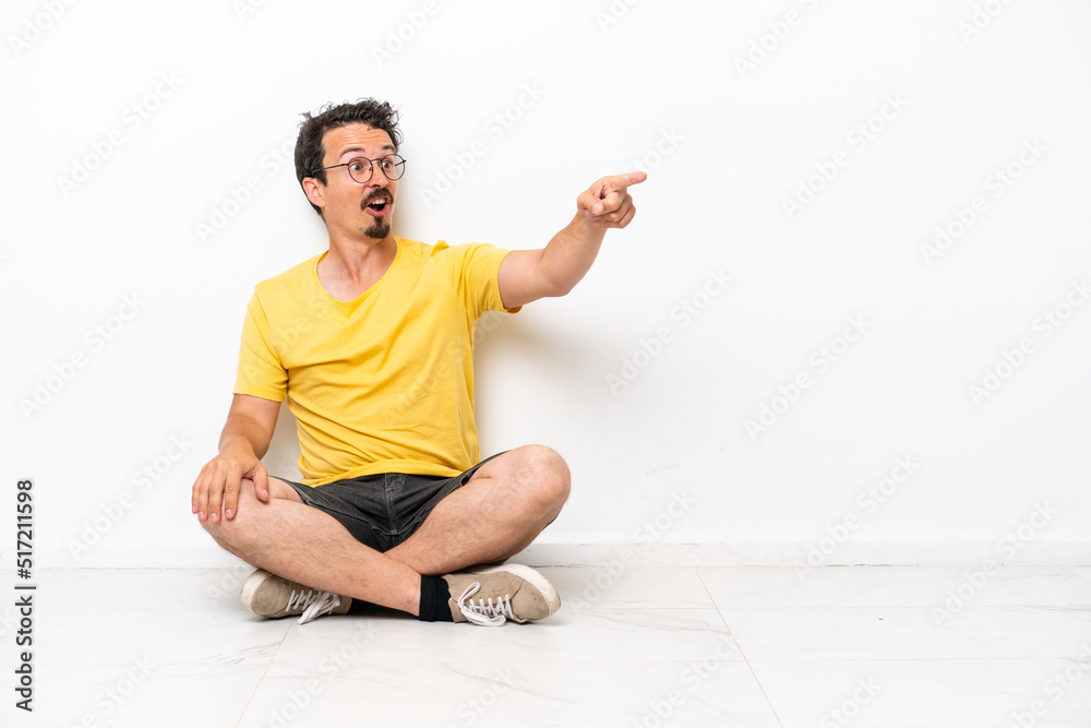 Fototapeta premium Young caucasian man sitting on the floor isolated on white background pointing away